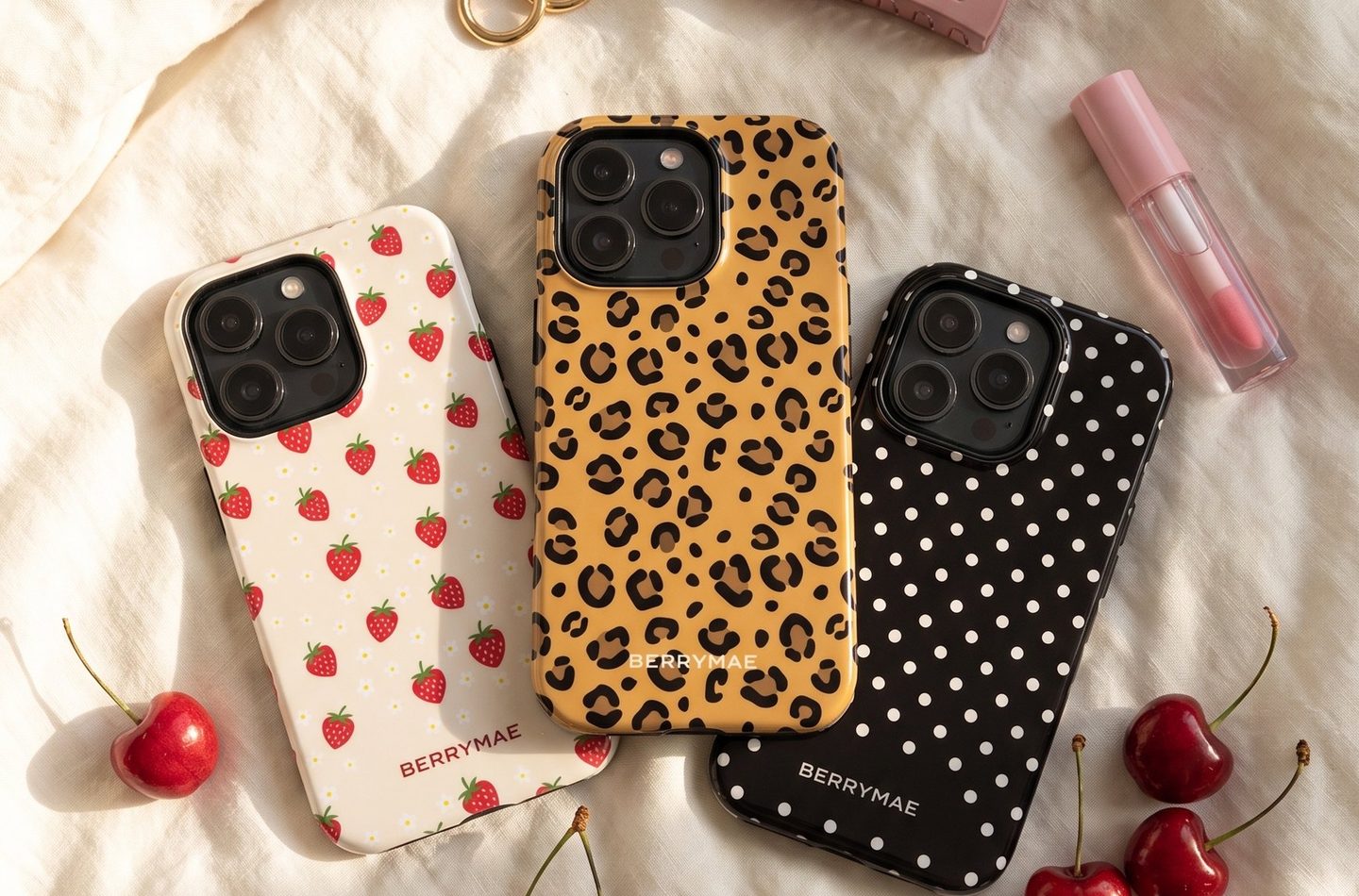 Three BerryMae phone cases on white linen, in strawberry, leopard and polka dot designs, with cherries beside them
