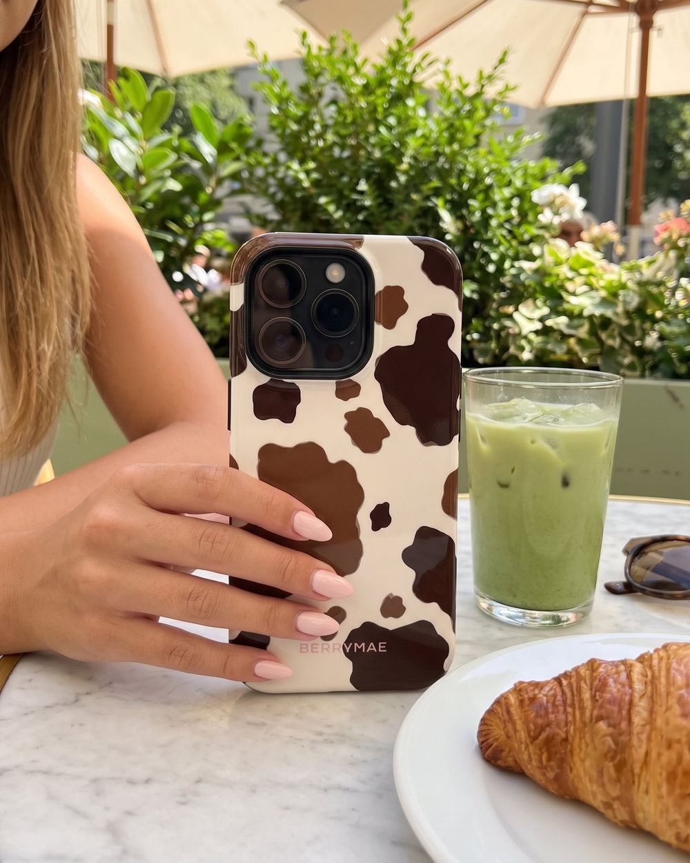 A hand holding a phone in a brown cow print BerryMae case at a cafe table, beside a croissant and an iced matcha